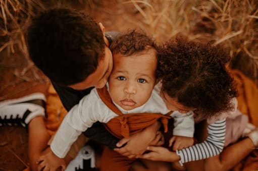 two older siblings cuddle and kiss their baby who is sitting on their lap in the spinifex grass. Baby stares up at the camera